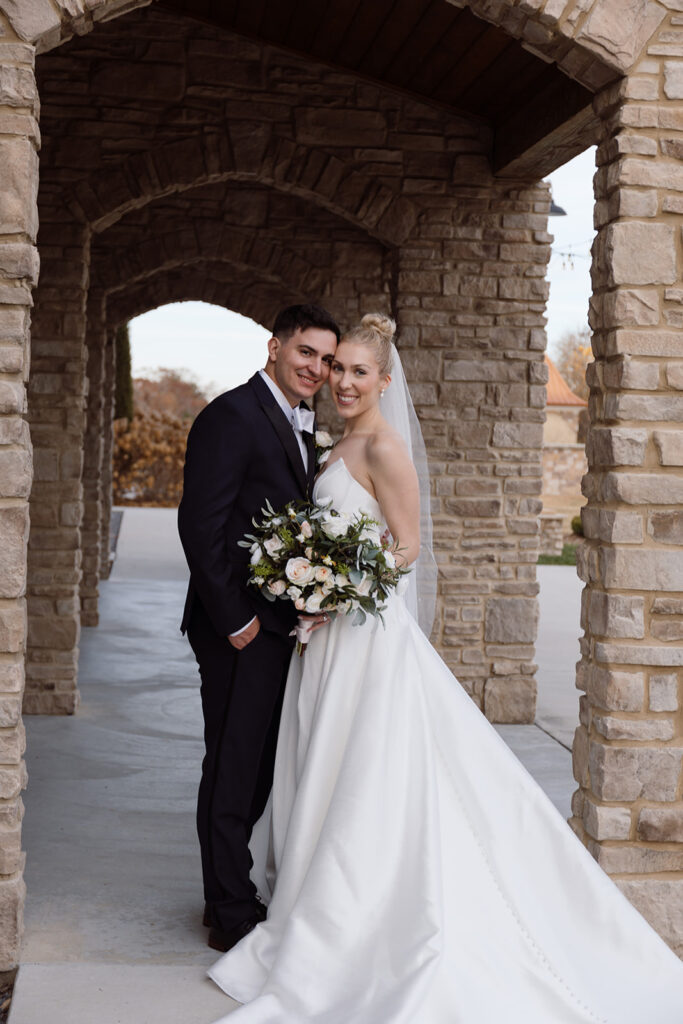 A newlywed couple poses together under a stone archway, smiling and holding a bouquet of flowers.