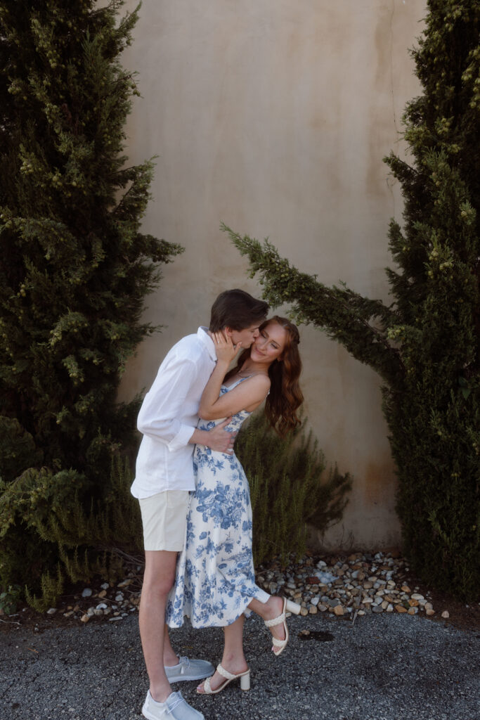 A couple embraces in a romantic pose against a textured wall, surrounded by greenery and stones, conveying a sense of intimacy and joy.