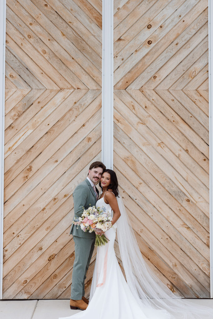A bride and groom pose together in front of a wooden chevron-patterned door, the bride holding a bouquet of flowers and wearing a sleek white gown, while the groom is dressed in a light green suit.