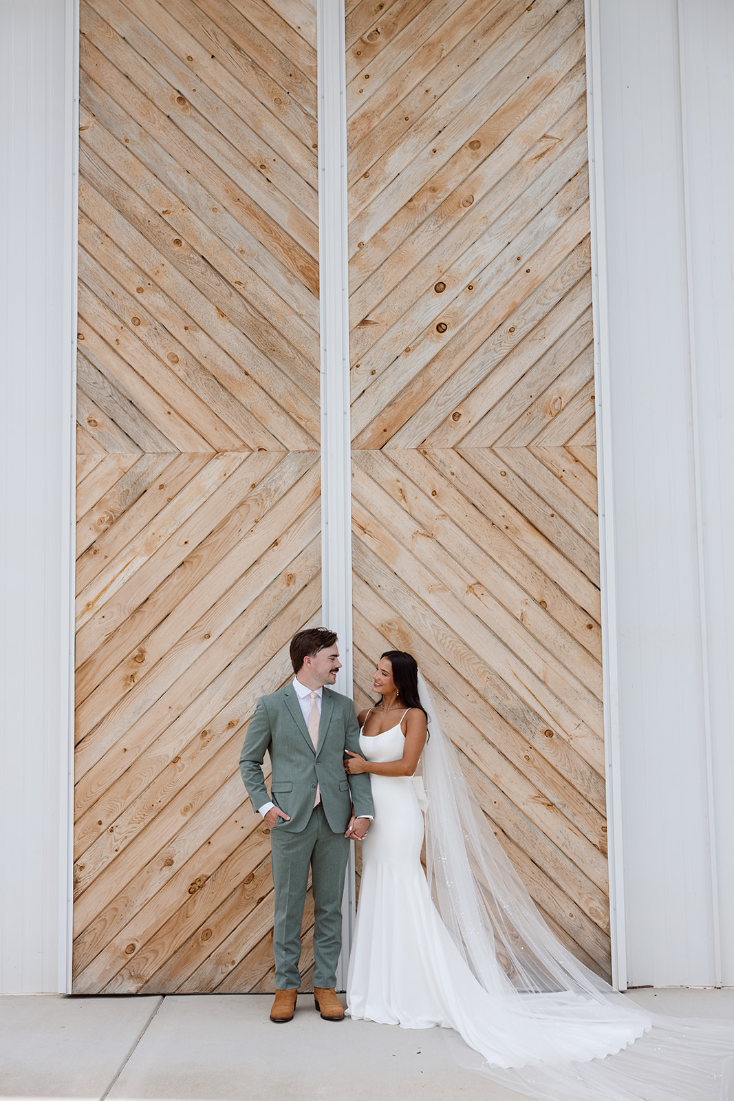 Bride and groom standing hand in hand in front of a large, modern wooden chevron wall. The bride wears a fitted white gown with a long train and veil, while the groom wears a sage green suit with a pink tie.