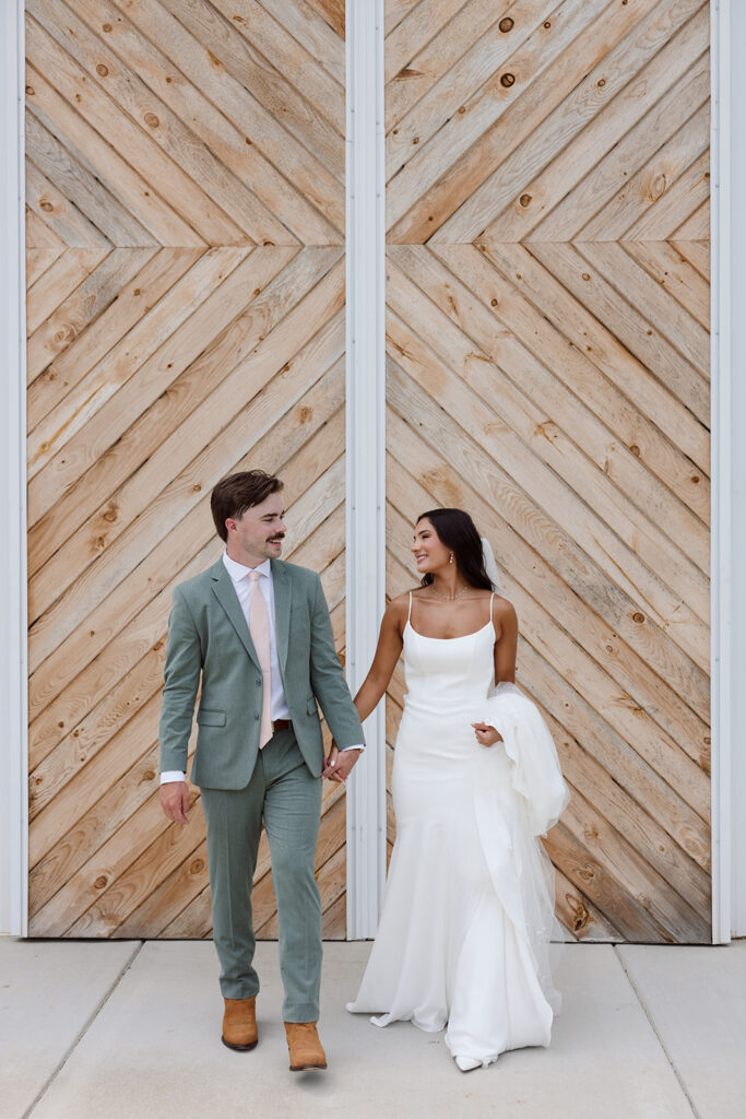 A bride and groom walking hand in hand in front of a rustic wooden wall, showcasing elegance and joy on their wedding day.