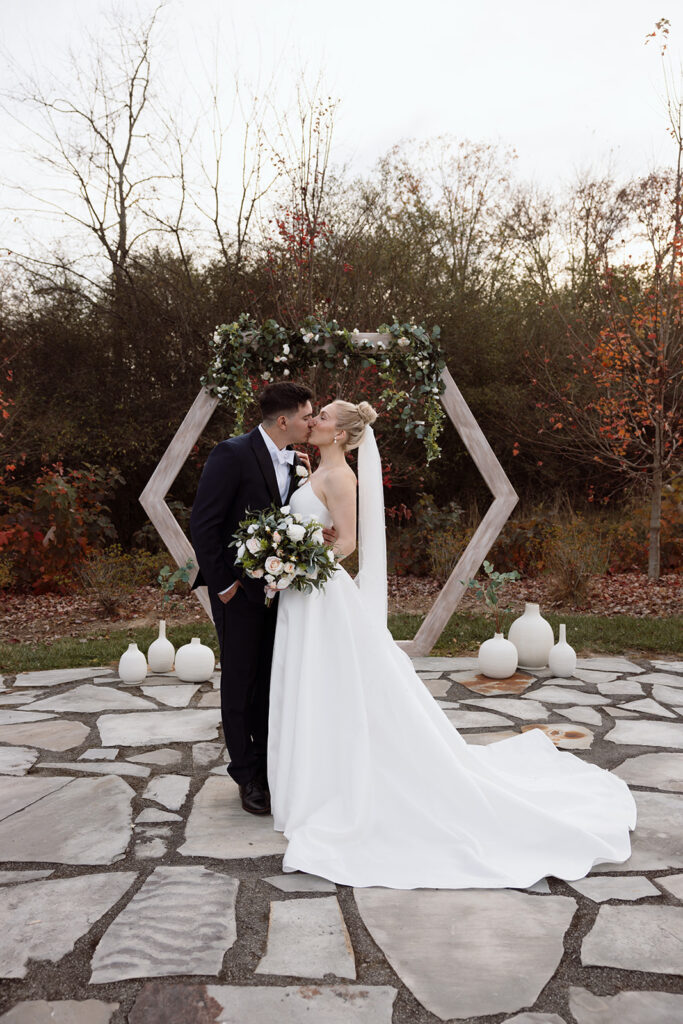 Couple kissing in front of a floral archway at an outdoor wedding ceremony on a stone patio.