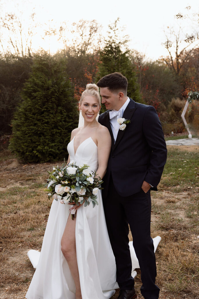 A bride in a white wedding dress holds a bouquet of flowers while posing with her partner, who is dressed in a black suit, in an outdoor setting with trees and a sunset backdrop.