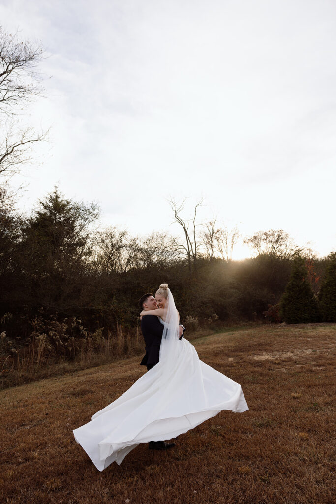 A joyful couple embracing on their wedding day, with the bride wearing a flowing white gown and veil, and the groom in a dark suit. The couple is surrounded by natural landscape with trees and an overcast sky.