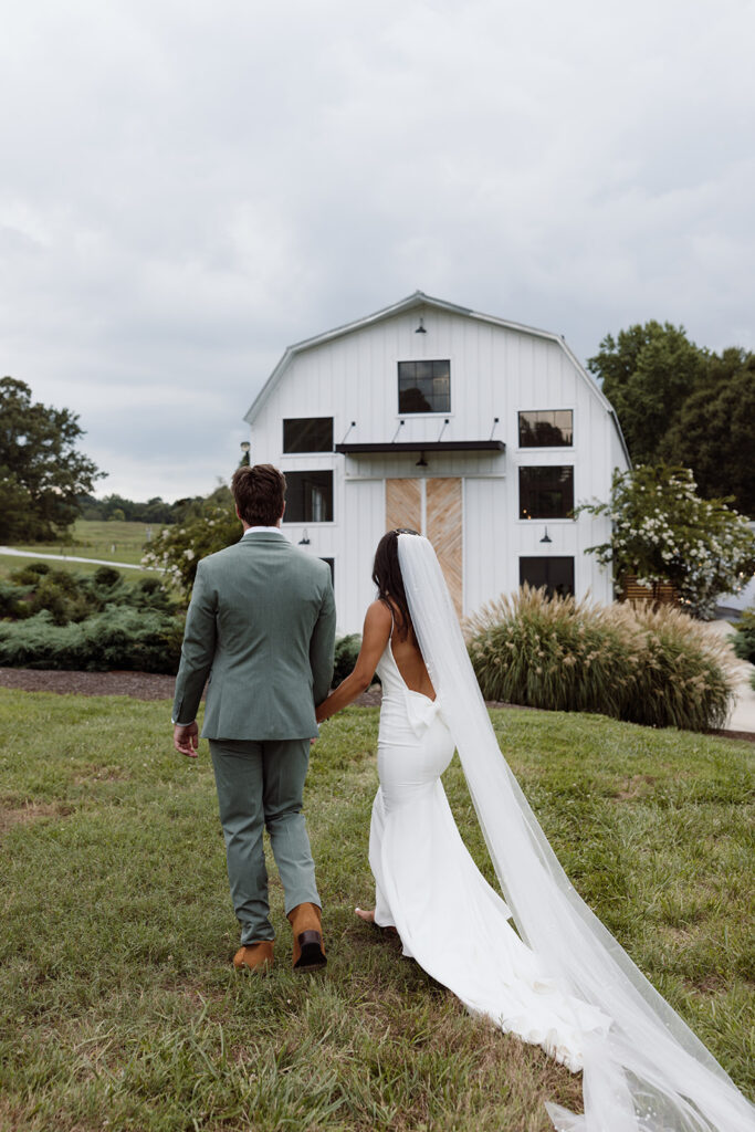 A couple walks hand-in-hand towards a modern white barn venue with a grassy foreground and cloudy sky, showcasing a romantic outdoor setting perfect for weddings.