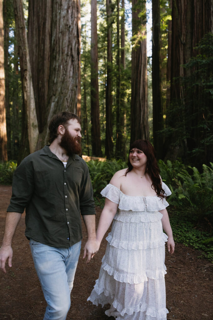 A couple walks hand in hand among towering redwood trees in a lush forest, smiling at each other.