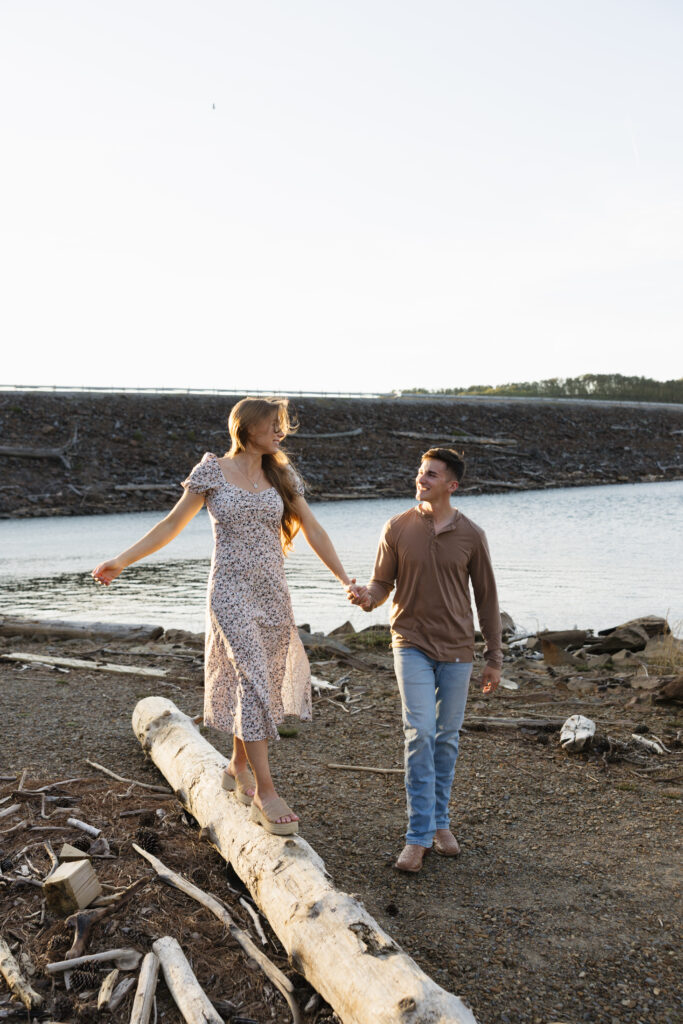 A couple joyfully walking hand-in-hand along a log by a lake, with the sun shining and a serene landscape in the background.