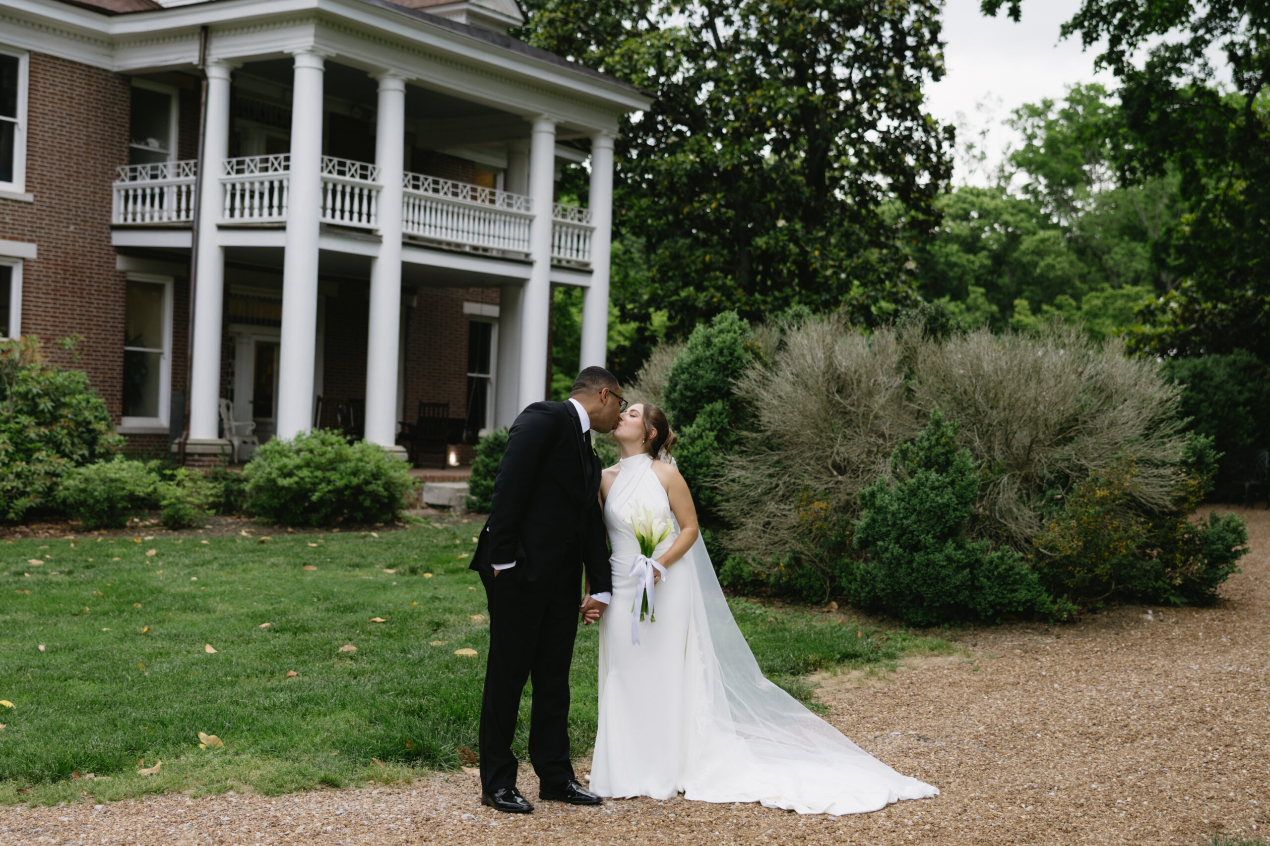 Bride and groom share a kiss in front of the historic brick mansion at Homestead Manor, surrounded by lush greenery.