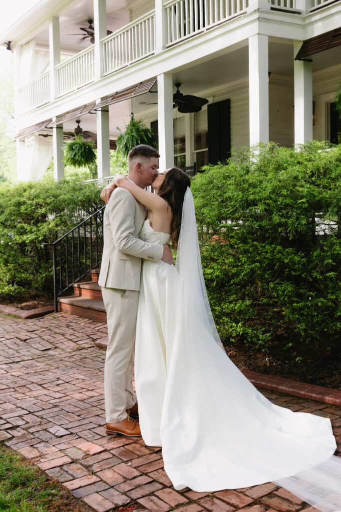 A couple sharing a kiss in front of a historic Southern-style home, surrounded by lush greenery and a brick pathway.