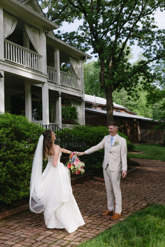 A bride in a flowing white gown holds hands with a groom in a light suit on a picturesque path surrounded by greenery and a charming Southern home.