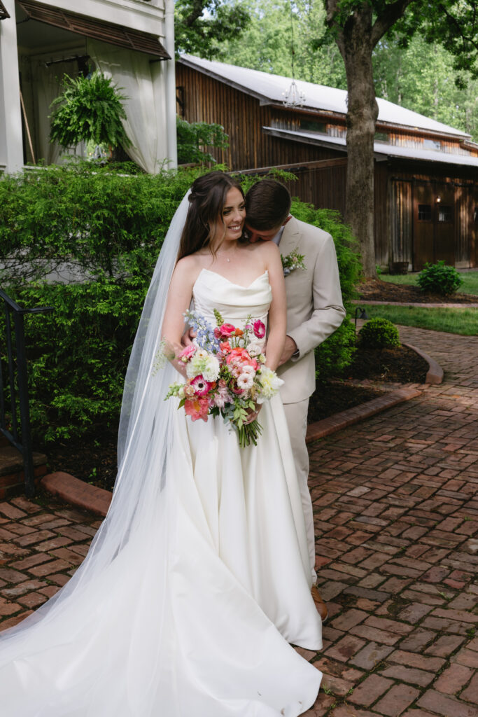 A bride in a strapless wedding gown holds a colorful bouquet while standing close to her partner, who is dressed in a light suit. They are outdoors, surrounded by lush greenery and rustic buildings, exuding a romantic and joyful atmosphere.