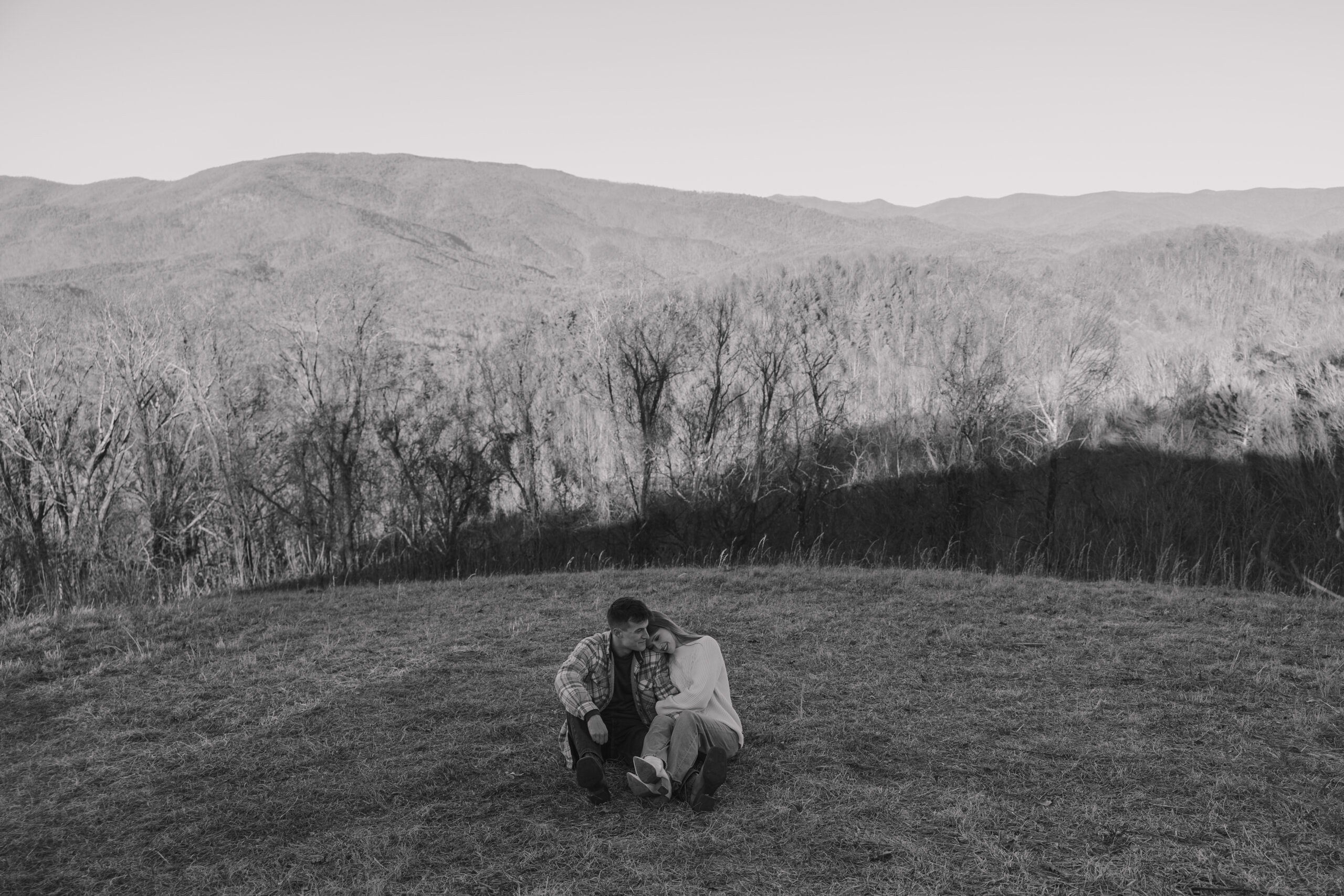 Couple sitting in a grassy field with mountains in the background, sharing a quiet, intimate moment during an engagement session.